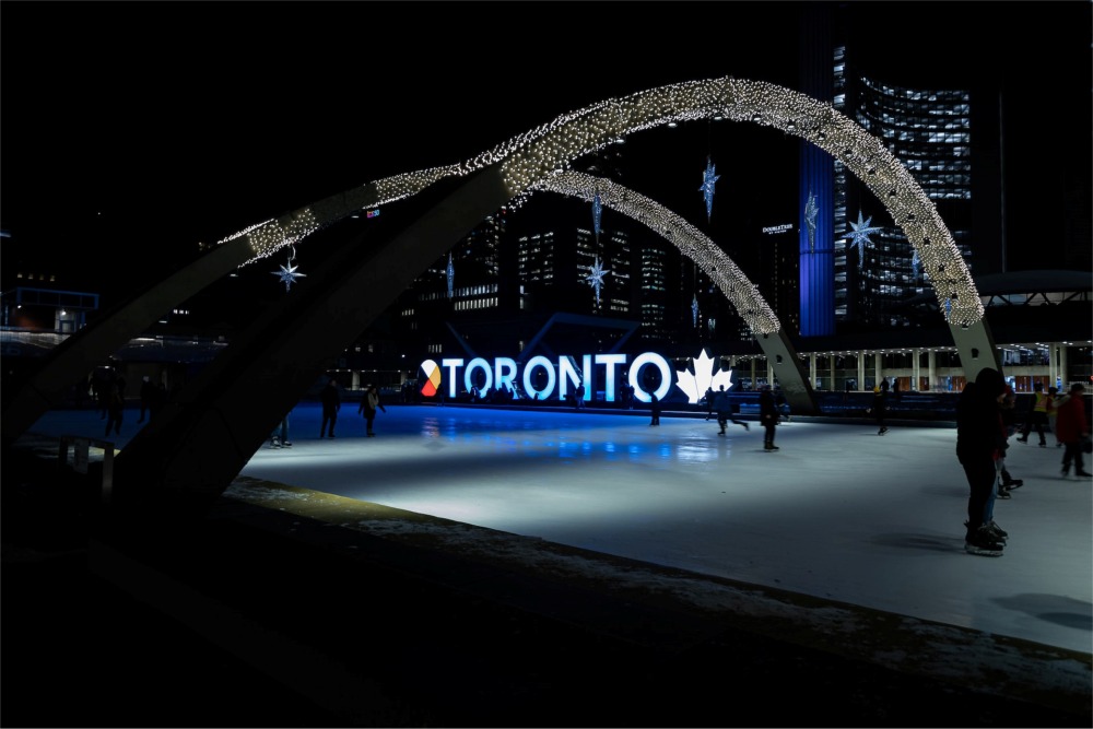This is a photograph of people ice skating at night in the Nathan Phillips Square in Toronto, Ontario, Canada. There is a nice view of the 3D Toronto sign in the background.