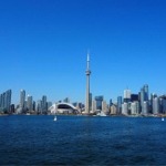 Toronto skyline with Lake Ontario in the foreground. It is a bright sunny day — Roger's Centre and the CN Tower feature in the middle of the image.