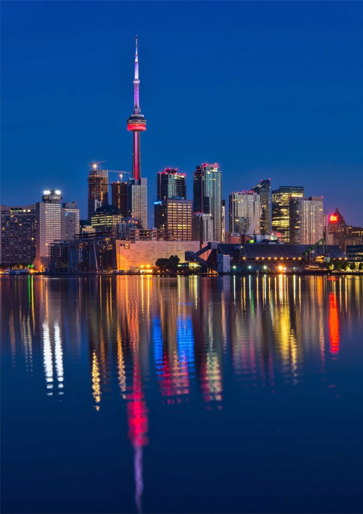 Photograph of the Toronto skyline at nighttime with Lake Ontario in the foreground. We see a lovely reflection of the colored city lights in Lake Ontario.