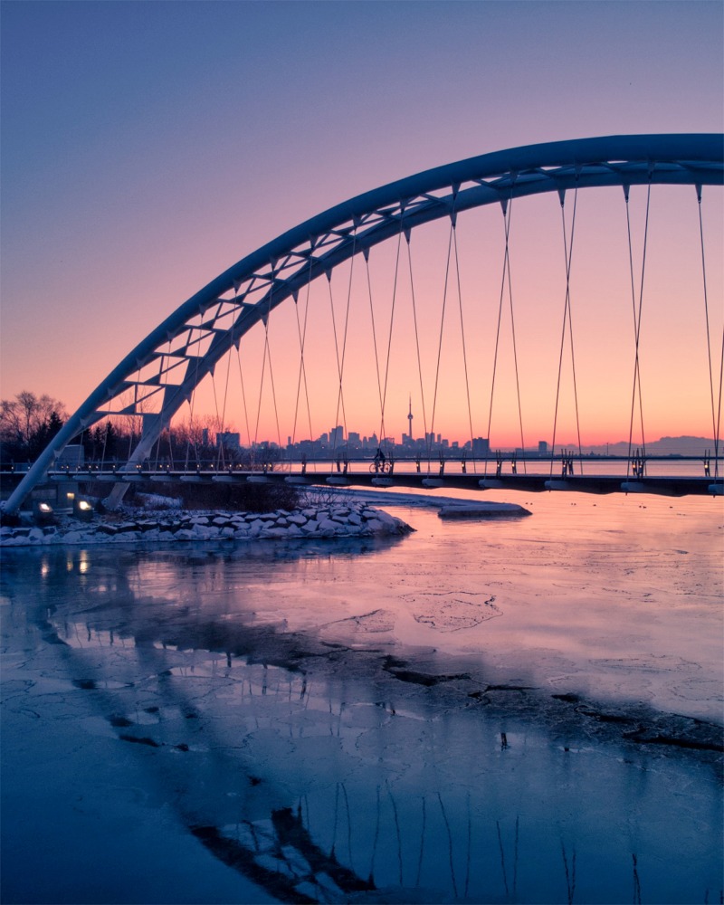 Photograph of the Humber Bay Arch Bridge in Toronto, Ontario, Canada. In the winter photo, we can see the CN tower through the arch and a cyclist on the bridge.